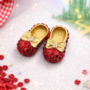 Puede incluir: Un par de zapatos rojos en miniatura con joyas, lazos dorados y una gema transparente en el centro. Los zapatos recuerdan a los icónicos zapatos de rubí. El fondo incluye un arcoíris y un castillo verde.