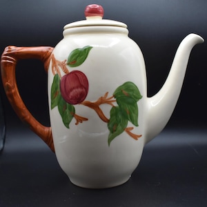 May include: Cream-colored ceramic teapot with a lid and a brown handle. The teapot is decorated with a red apple and green leaves on a branch. The handle has a bamboo-like design.