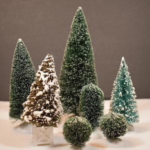 Puede incluir: Una colección de árboles de Navidad en miniatura de cepillo para botellas en varios tonos de verde y marrón, algunos cubiertos de nieve blanca. Los árboles varían en tamaño y forma, creando una exhibición festiva.