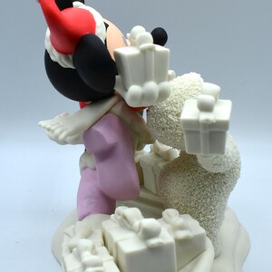 Op de afbeelding: Een beeldje met een stripfiguur met een kerstmuts, zwarte oren en roze kleding, met witte cadeau dozen. Er is ook een sneeuwpopfiguur aanwezig, met cadeaus. De basis is wit met meer cadeau dozen.
