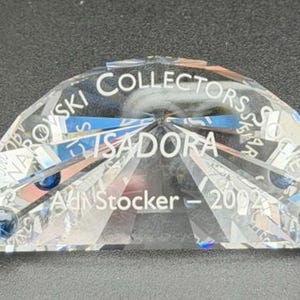 May include: A clear crystal collectible with the text "Collectors Society Isadora" and "A.N. Stocker - 2002". The crystal has a half-circle shape with facets and two blue crystal accents. The collectible is on a dark surface.