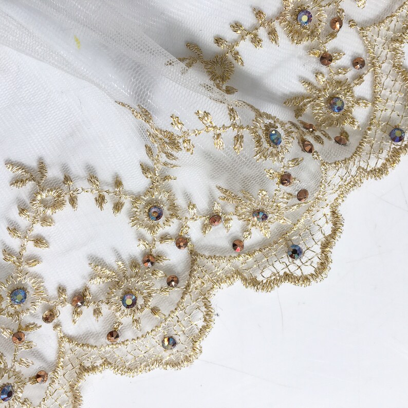 May include: Gold lace trim with iridescent beads and a scalloped edge. The lace is on a white background.