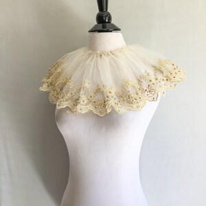 May include: A white lace collar with gold embroidery and rhinestones. The collar is ruffled and has a scalloped edge.