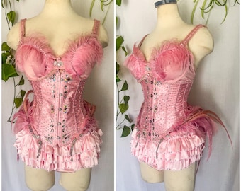 May include: A pink satin corset with a bustier top and a ruffled skirt. The corset is decorated with rhinestones and feathers.