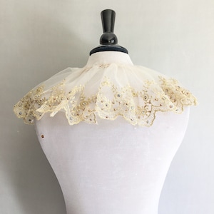 May include: A white lace collar with gold embroidery and clear rhinestones. The collar is on a white dress form.