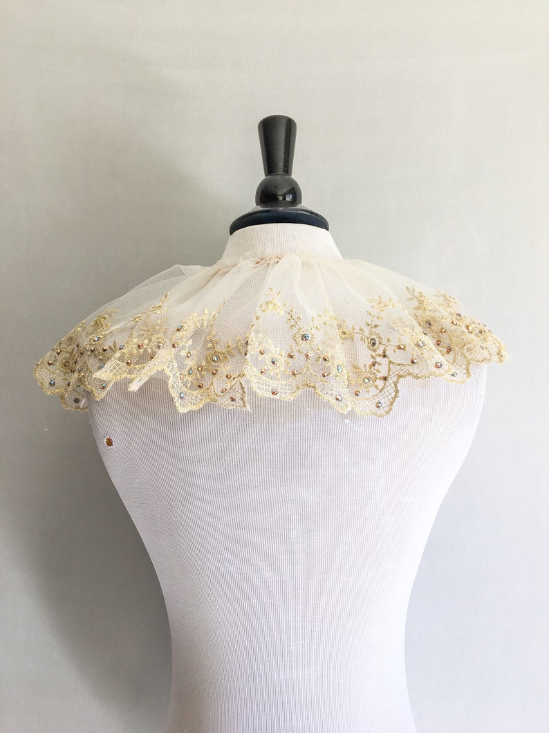 May include: A white lace collar with gold embroidery and clear rhinestones. The collar is on a white dress form.
