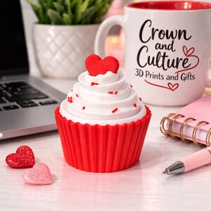 May include: A red and white cupcake-shaped item with a heart on top, red sprinkles, and a red cupcake liner. A white mug with the words "Crown and Culture" and "3D Prints and Gifts" is in the background. Two heart-shaped decorations are in the foreground.