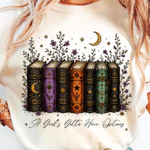 Könnte beinhalten: Ein cremefarbenes Sweatshirt mit einer Grafik aus sieben Vintage-Büchern mit goldenen Verzierungen, Sternen und Mondsicheln. Über den Büchern befinden sich violette Blumen und ein Mond. Der Text lautet, A Girl's Gotta Have Options.