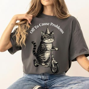 May include: A charcoal gray t-shirt featuring a graphic of a tabby cat wearing a party hat and riding a scooter. The text above the cat reads "Off To Cause Problems" in white lettering. The cat has a serious expression and striped fur.