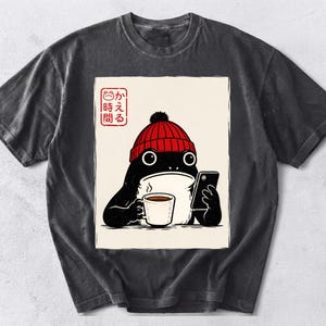 Puede incluir: Una camiseta gris carbón con el gráfico de una rana negra que lleva un gorro rojo, sosteniendo una taza de café blanca humeante y un teléfono inteligente negro, con texto en japonés y un pequeño icono de rana en la esquina superior izquierda.