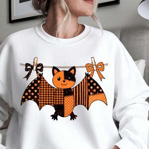 May include: A white crewneck sweatshirt featuring a graphic of a bat hanging from a clothesline with clothespins and bows. The bat has a patchwork design in orange and black, incorporating patterns like polka dots, stripes, and plaid.