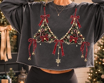May include: A charcoal grey cropped sweatshirt featuring a graphic print of two triangular Christmas tree shapes decorated with red velvet bows, candy canes, pinecones, gold stars, string lights, and hanging gold bells and a snowflake charm.