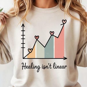 Może przedstawiać: Beżowa bluza z nadrukiem wykresu z sercami i napisem "Healing isn't linear". Wykres przedstawia nieliniowy przebieg z kolorowymi słupkami i ikonami serc, symbolizującymi dobre samopoczucie emocjonalne.