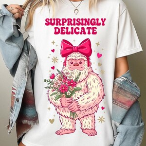 Könnte beinhalten: Ein weißes T-Shirt mit einer Grafik eines hellrosa Yetis, der eine große, kräftig rosa Schleife auf dem Kopf trägt. Der Yeti hält einen Strauß bunter Blumen. Über dem Yeti steht der Text "SURPRISINGLY DELICATE" in kräftigen, rosa Buchstaben.
