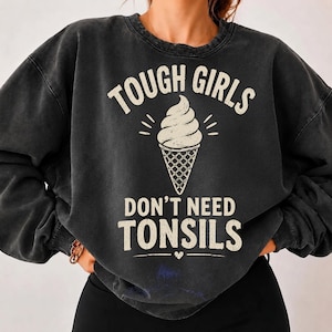 May include: A charcoal black crewneck sweatshirt with a vintage-style wash. The front features a graphic of a soft-serve ice cream cone in cream colour, with the text "TOUGH GIRLS DON'T NEED TONSILS" printed in a distressed, bold, sans-serif font.
