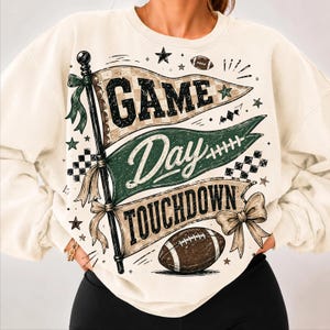 Puede incluir: Una sudadera de cuello redondo color crema con un estampado gráfico de tres banderines apilados en un mástil. Las banderas muestran el texto "GAME DAY" y "TOUCHDOWN" en negro y verde, acentuado con balones de fútbol, estrellas y lazos decorativos.