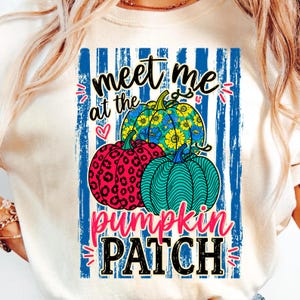 May include: A cream-colored t-shirt featuring a graphic print of three pumpkins in front of blue vertical stripes. The pumpkins have floral, leopard print, and wavy line patterns. The text reads "meet me at the pumpkin PATCH" in black and pink.