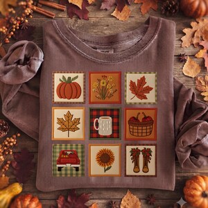 Könnte beinhalten: Ein braunes Sweatshirt mit Rundhalsausschnitt und einem Raster aus neun quadratischen Illustrationen. Die Grafiken zeigen einen Kürbis, Wildblumen, Ahornblätter, eine Kaffeetasse auf rotem Karomuster, einen Korb mit Kürbissen, einen roten Lastwagen, eine Sonnenblume und Stiefel.