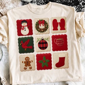 May include: A cream-colored t-shirt featuring a grid of nine square patches with Christmas-themed designs, including a snowman, a wreath, red mittens, a cardinal, a buffalo plaid ornament, a mug, a gingerbread man, holly, and a stocking.