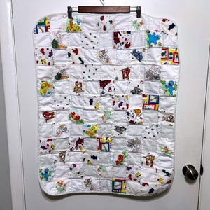 May include: White patchwork baby quilt with various cartoon character squares. The quilt is hanging on a wooden hanger. The squares feature colorful images of animals and characters.