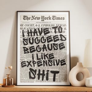 May include: A wooden media unit with a ribbed front, displaying a framed print from The New York Times with the text "I HAVE TO SUCCEED BECAUSE I LIKE EXPENSIVE SHIT." A lamp, vase, and stereo are also on the unit.