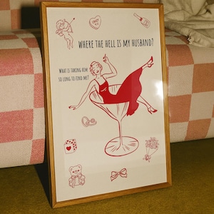 May include: Framed print with a red illustration on a white background. The artwork features a woman in a martini glass, surrounded by small drawings of a cupid, dice, and a teddy bear. Text reads "WHERE THE HELL IS MY HUSBAND?" and "WHAT IS TAKING HIM SO LONG TO FIND ME?"