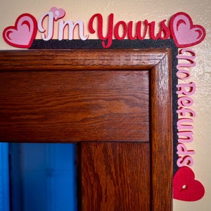 May include: A wooden door frame with a decorative border. The border features the text "I'm Yours" in red and white, along with pink and red heart shapes. The words "no refunds" are also visible.