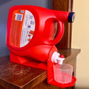 May include: A red laundry detergent bottle with a dispenser and measuring cup. The bottle has a handle and a blue cap. The dispenser is white with a red stopper. The measuring cup is clear plastic.