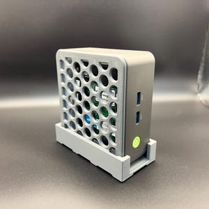 May include: A gray computer case with a perforated front panel and a matching stand. The case has a rounded top and a series of ports on the side, including USB ports. A green power button is visible.