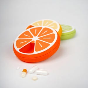 Könnte beinhalten: Drei farbenfrohe Pillenboxen in Form von Zitrusfruchtscheiben: Orange, Zitrone und Limette. Der orangefarbene Organizer ist geöffnet und zeigt ein Fach für Pillen. Mehrere Pillen und Kapseln liegen vor den Organisatoren verstreut.