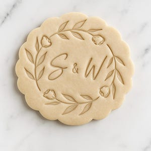 May include: A round biscuit with a scalloped edge, featuring the initials "S & W" in elegant script, surrounded by a floral wreath design. The biscuit is a light tan colour, with the floral and initial details slightly darker. The biscuit is a wedding or engagement themed item.