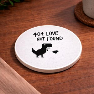 May include: White ceramic coaster with the text "404 LOVE NOT FOUND" and a pixelated dinosaur graphic. The coaster has a round shape and a slightly textured surface. A small heart graphic is also present.