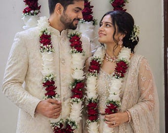 May include: A couple in cream-colored wedding attire, adorned with floral garlands of white roses and deep red flowers. The garlands are draped around their necks, complementing their outfits. The background features similar floral decorations.