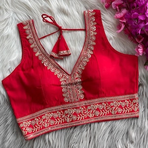 May include: A sleeveless red blouse with a deep V-neckline. The blouse is adorned with intricate gold embroidery along the neckline, armholes, and hem. A matching tassel detail hangs from the back.