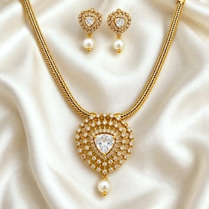 May include: A gold-toned jewelry set featuring a necklace with a thick braided chain and a circular pendant containing a central triangular clear stone surrounded by small white crystals and a dangling pearl. Matching triangular earrings included.