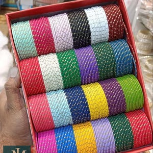 Pode incluir: Uma caixa vermelha cheia de várias pilhas de pulseiras coloridas. As pulseiras são em várias cores, incluindo rosa, azul, verde, amarelo e roxo, com detalhes em pontos dourados. O logotipo "jewelindiamart" é visível.