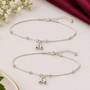 May include: A pair of silver-toned anklets featuring a delicate chain with evenly spaced small beads. Each anklet is finished with a lobster claw clasp, an adjustable extender chain, and a central charm consisting of two small, round silver bells.