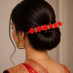 May include: A hair accessory featuring a row of five bright red fabric flowers with small green leaf accents on a thin gold-toned metal bar. The accessory is secured horizontally across a dark brown hair bun, complemented by gold-toned jhumka earrings.