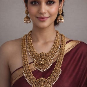 May include: A gold-toned jewelry set featuring a two-layered necklace and matching jhumka earrings with a matte finish. The necklace displays intricate circular motifs and central deity pendants, accented with small white beads. Text reads No.2010-515.