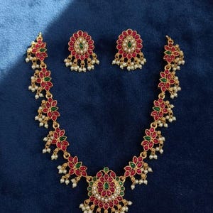 May include: A gold-toned necklace and earring set with red and green floral accents and small white pearl-like beads. The earrings and necklace feature a matte finish and are displayed on a dark blue background. The text "No906-230 Matte Finish Premium Quality" is visible.