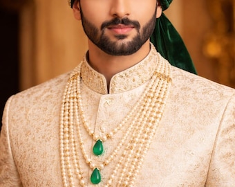 May include: A man wearing a cream-colored embroidered jacket and a green turban. The jacket is paired with a multi-strand pearl necklace featuring emerald green pendants. The turban is adorned with gold embellishments.