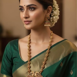 May include: A long gold necklace featuring spherical beads and a large, ornate pendant with green and red stones, paired with matching jhumka earrings. The jewelry is worn with a dark green saree with a gold border. Text reads No.1700-425 Matte Finish Priemum Quality.