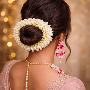 Pode incluir: Um coque adornado com uma guirlanda floral branca circular, combinado com brincos pendentes de flores rosas e vermelhas e um colar dourado com um fio vertical de pérolas brancas, usado sobre uma peça de roupa rosa bordada com detalhes dourados.