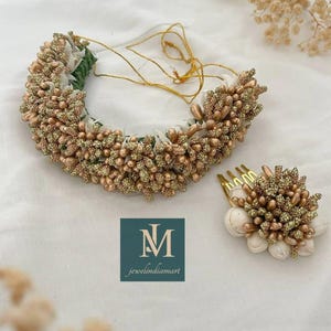 Op de afbeelding: Een bloemenhaarkrans en een bijpassende haarkam met kleine witte stoffen bloemen en metallic goudkleurige kralen met een glitterafwerking. De set bevat dunne gouden koordjes en een logo met de tekst M jewelindiamart.