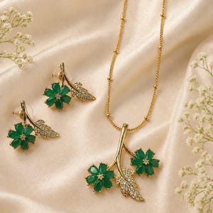 May include: A gold-coloured jewellery set comprising a necklace and earrings. The necklace features a pendant with two green flower-shaped gems and a leaf with clear crystals. The earrings echo the pendant design. The set is displayed on a cream satin surface.