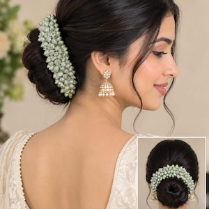 May include: A hair accessory featuring a cluster of pale green, bud-shaped beads arranged in a curved, elongated form. The accessory is pinned to a dark hair bun. Also included are gold-toned jhumka earrings with small white pearl-like beads.