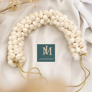 Peut inclure: Un accessoire de cheveux floral blanc, probablement une guirlande ou un chignon, en forme d'arche. Il est composé de petites fleurs blanches serrées. Une ficelle dorée est attachée aux deux extrémités. Le texte "jewelindiamart" est visible.