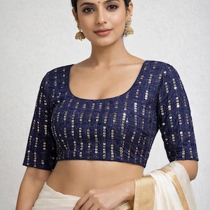 Puede incluir: Una blusa azul marino con detalles de lentejuelas doradas. La blusa tiene un escote redondo y mangas tres cuartos. La modelo lleva pendientes dorados y un sari blanco con borde dorado.