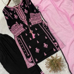 May include: A black long-sleeved tunic featuring intricate pink embroidery and mirror accents along the neckline, cuffs, and hem. The set includes matching black trousers with embroidered hems and a sheer, light pink dupatta with a black lace border.