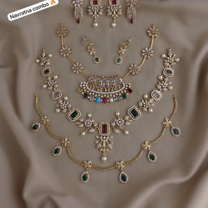 Può includere: Una collezione di parure di gioielli color oro, tra cui collane e orecchini, adornate con varie pietre preziose colorate e cristalli trasparenti. I set sono disposti su un tessuto beige. Il testo "Navratna combo" è visibile.